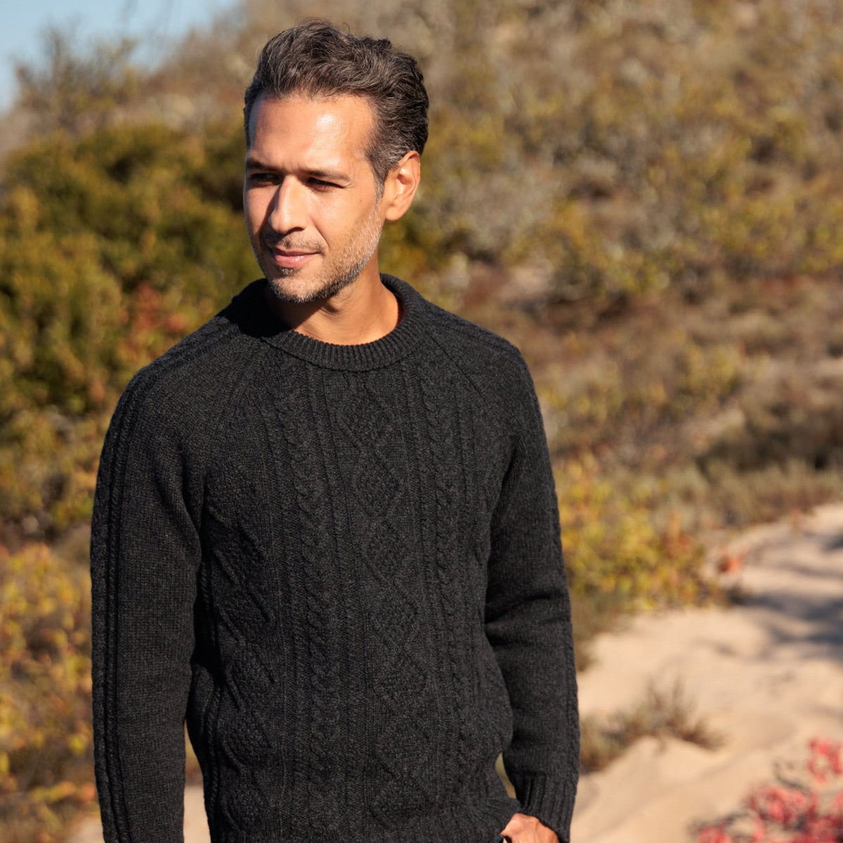 The Ross 4-ply Scottish lambswool cable knit crewneck in Charcoal, walking on the dunes